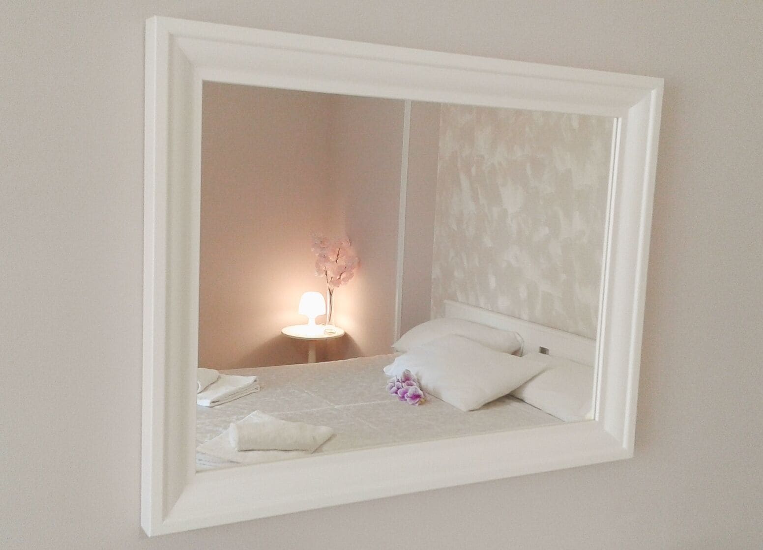 Un lit bien fait, avec des oreillers et une serviette pliée, une table de chevet avec une lampe, et un miroir fixé au mur qui reflète la pièce.
