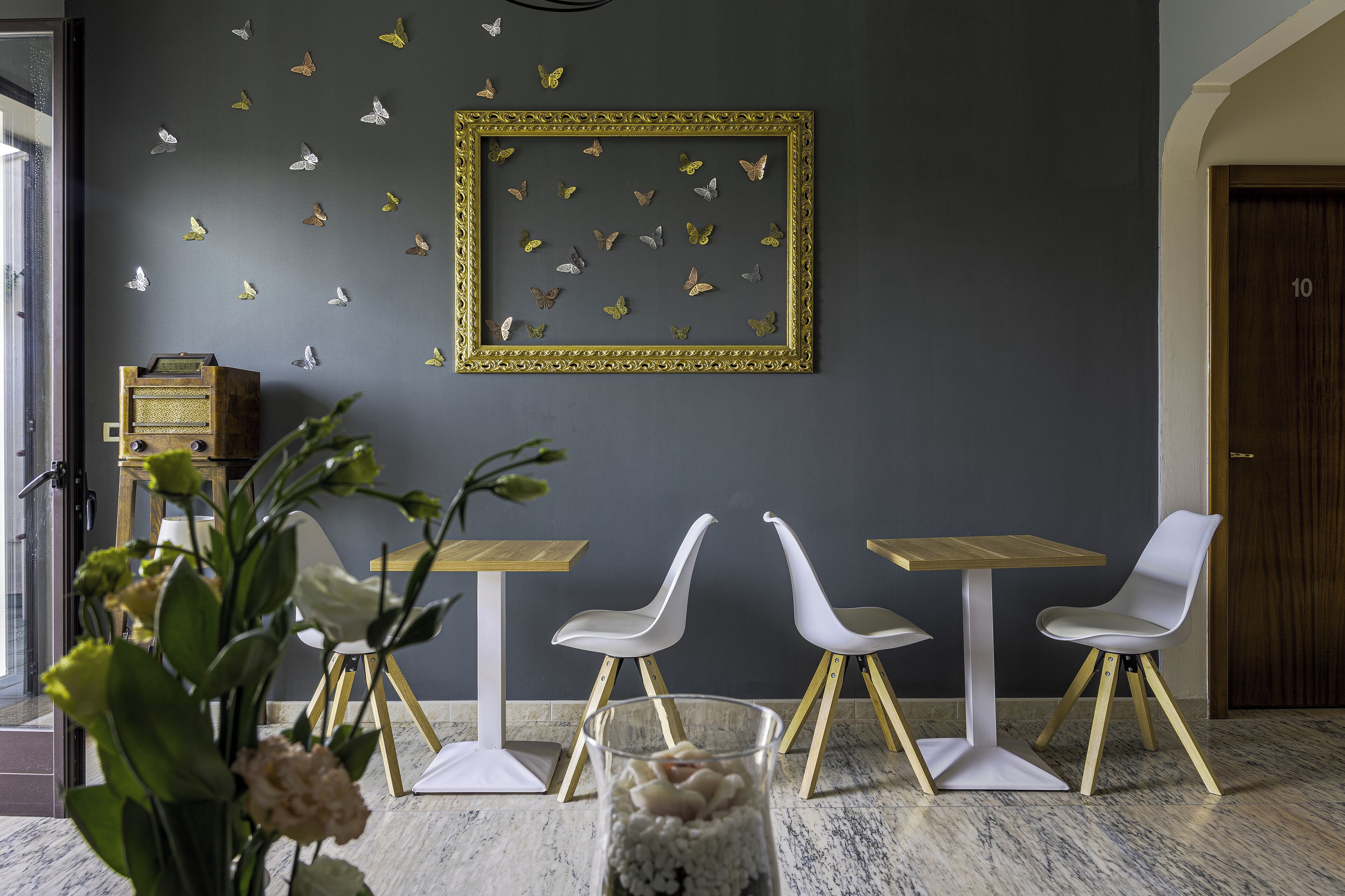 Un intérieur moderne avec un mur foncé, un tableau encadré représentant des papillons, des chaises blanches et une table en bois.