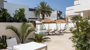 A poolside area with a white table and chairs, lounge chairs, and thatched umbrellas. - La Gemma del Salento (Racale)