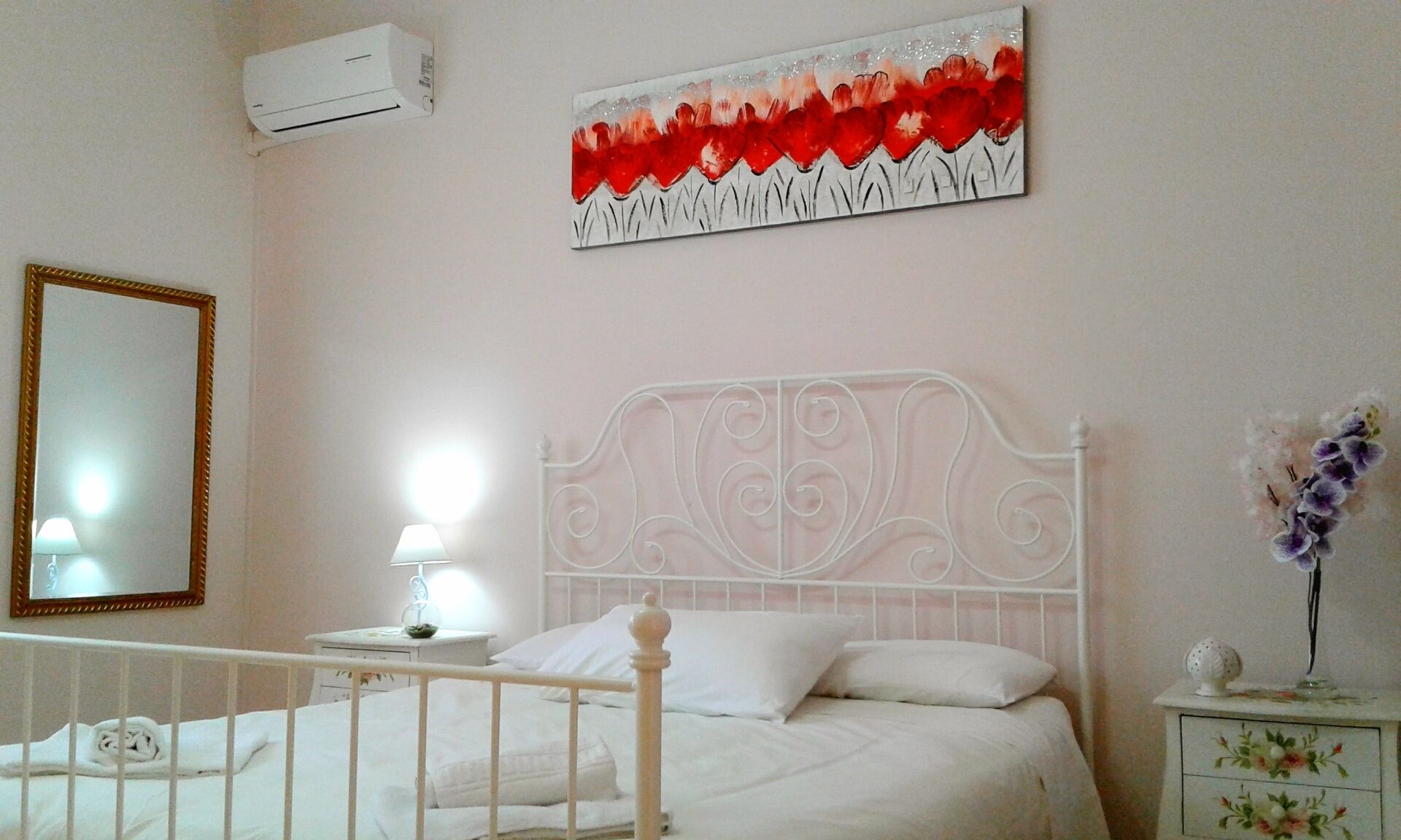 Une chambre avec un lit blanc, une table de chevet avec une lampe, un climatiseur fixé au mur et une œuvre d’art encadrée au-dessus du lit.