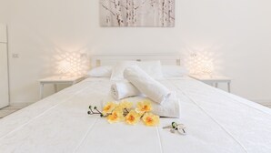 A neatly made bed with white linens and rolled towels, a painting of trees on the wall, and a small table with a lamp and a key on the bedside. - La Gemma del Salento (Racale)
