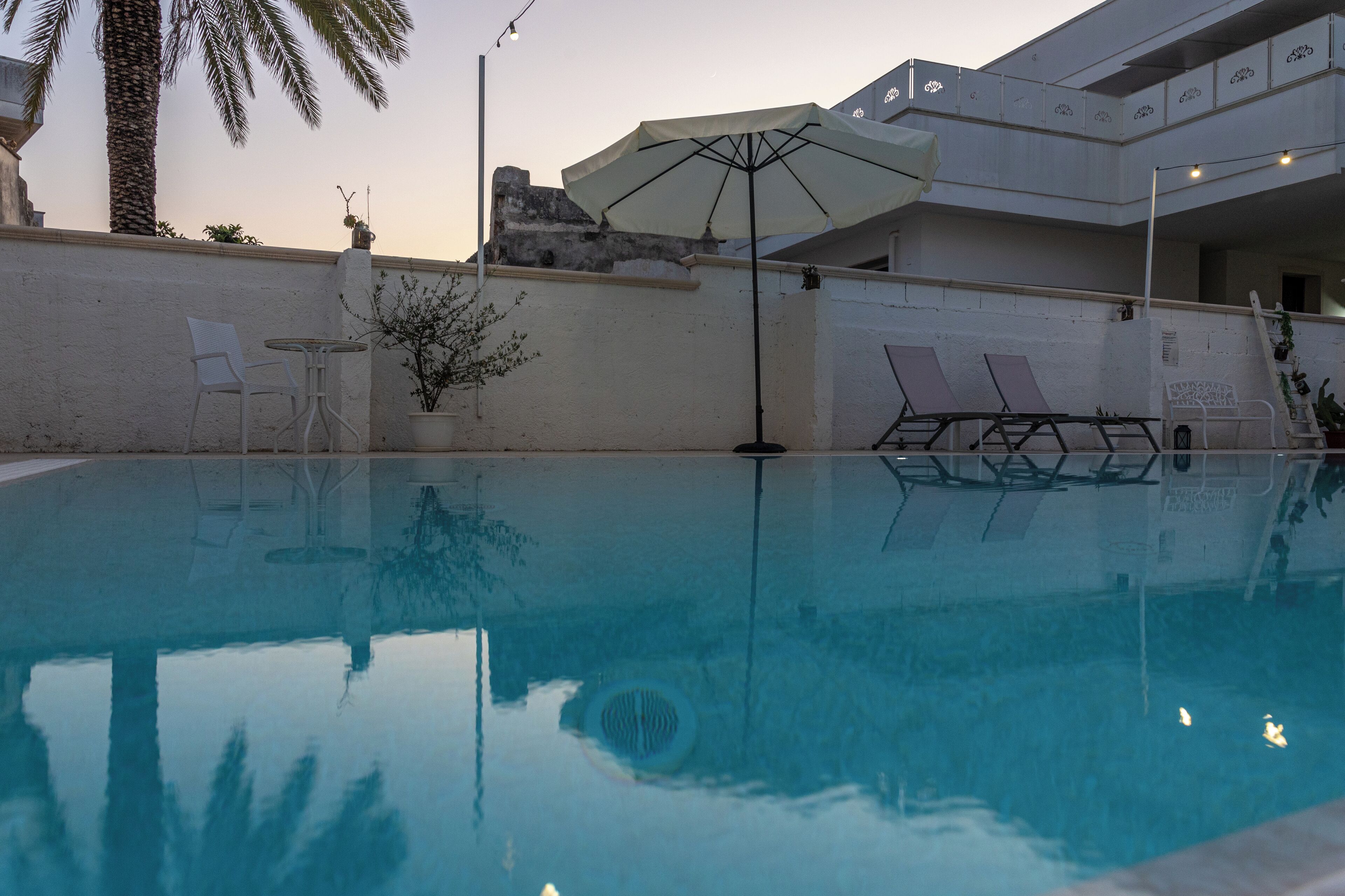 A swimming pool with lounge chairs and an umbrella, surrounded by a white wall and palm trees.