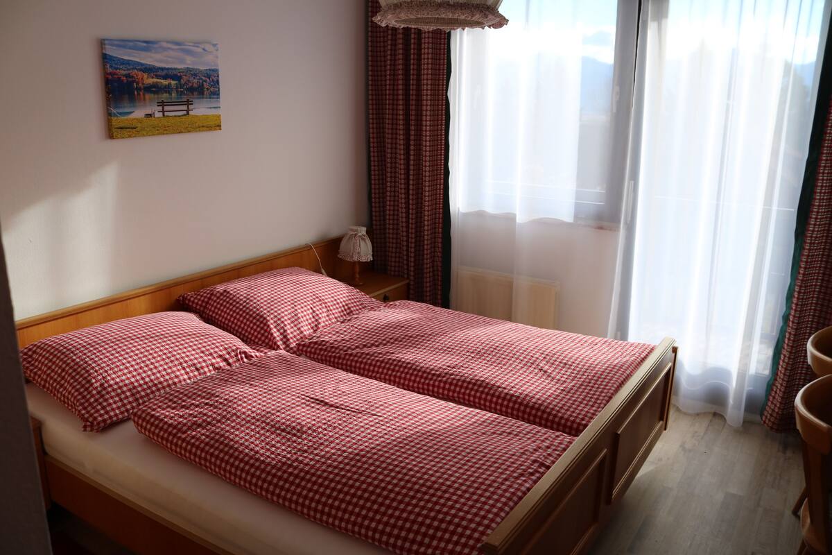 A double bed with checkered bedding, a wooden headboard, a nightstand with a lamp, a painting on the wall, and a window with curtains.