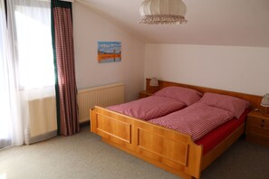 Quarto com cama de madeira, mesa de cabeceira, luminária, radiador, janela com cortinas leves e um quadro com paisagem na parede.