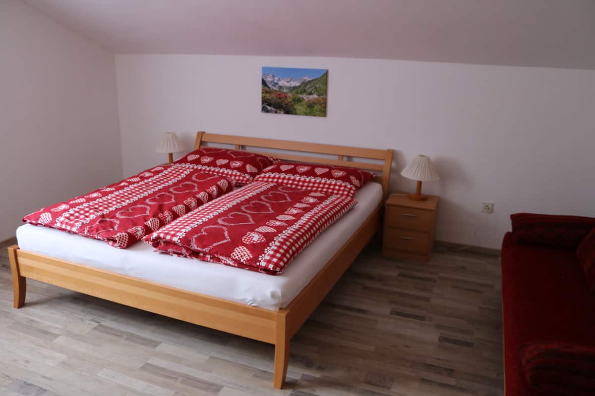 A bedroom with a wooden bed, a red and white patterned bedspread, two bedside lamps, a wooden nightstand, and a red sofa.