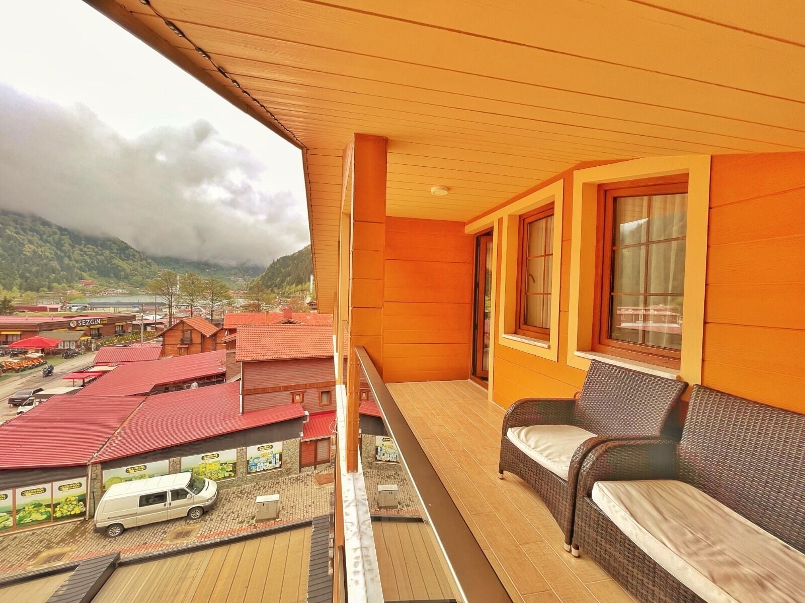 A balcony with a wicker sofa and a view of a town with red-roofed buildings and a lake.