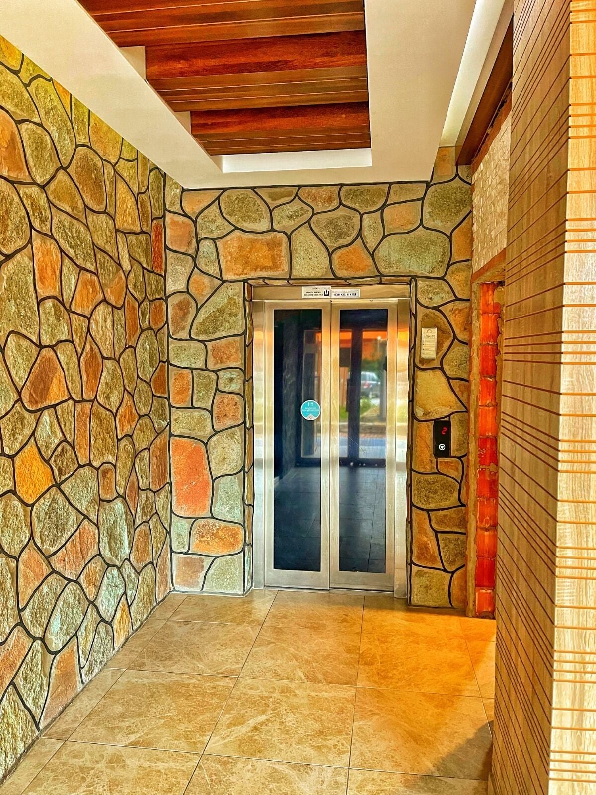 A stone-walled elevator with a glass door, a tiled floor, and a wooden ceiling.