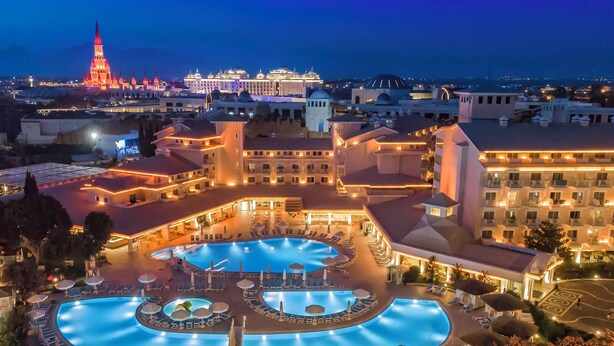 Un hotel con una amplia piscina, varias zonas de descanso y un edificio imponente al fondo.