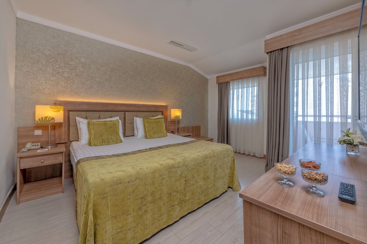A hotel room with a large bed, bedside tables with lamps, a wooden desk with a remote control, and a vase with flowers.