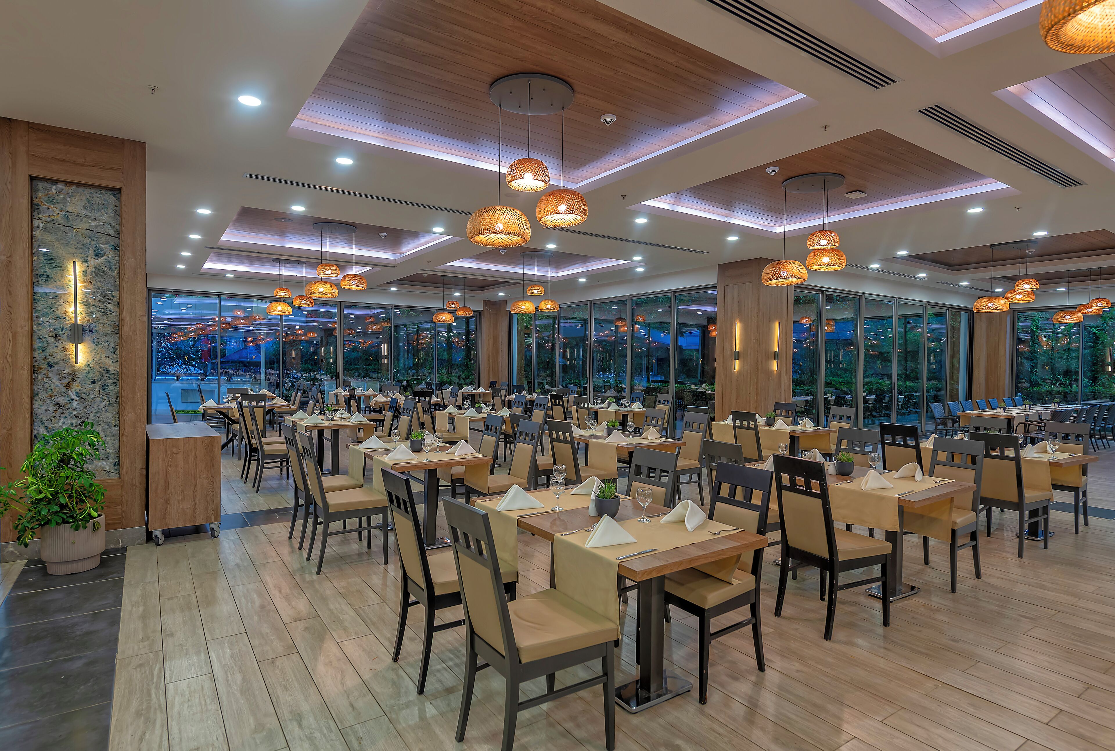 Un restaurant spacieux, doté de nombreuses tables et chaises, de grandes fenêtres offrant une vue sur le paysage urbain et d’un éclairage moderne suspendu au plafond.