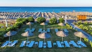 Un complejo turístico en la playa con filas de tumbonas y sombrillas de paja, una zona de piscina y vistas al mar.