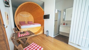 Een knus ogende kamer met een houten bed, een klein tafeltje met geruit tafelkleed, een douche en een witte wastafel.