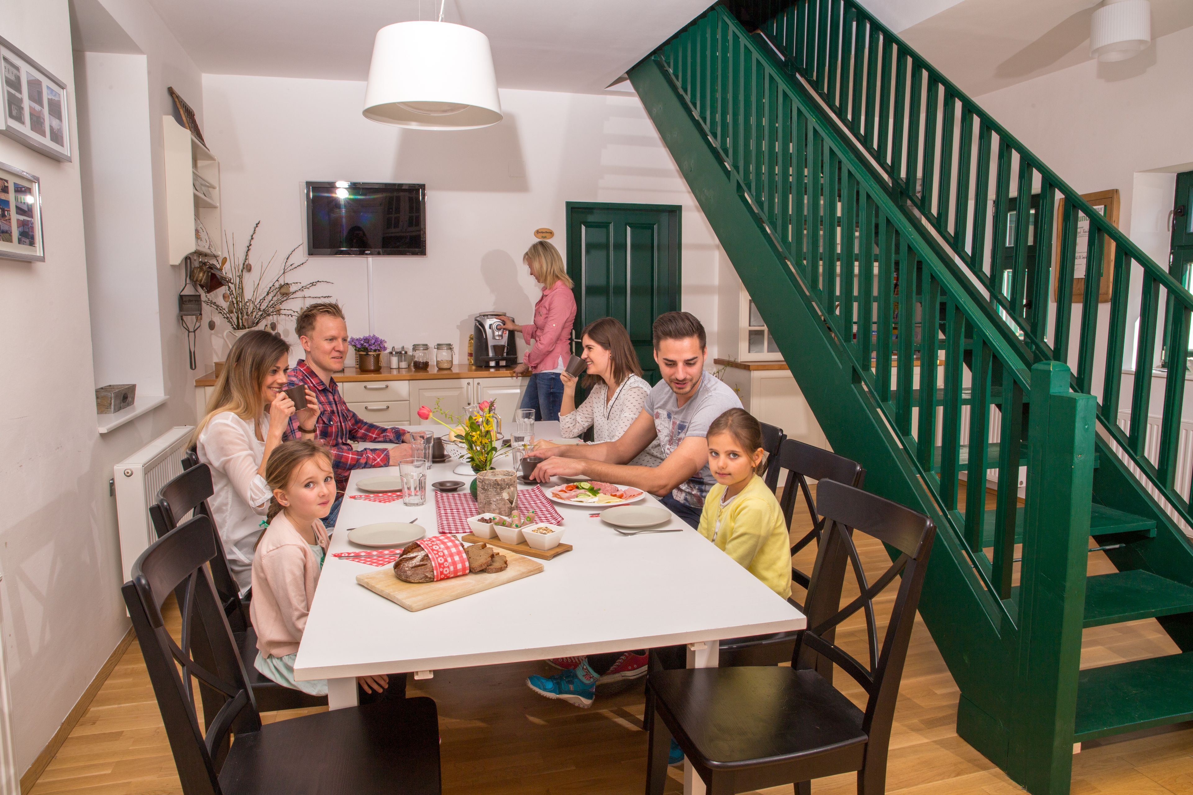 Una familia cenando en una mesa blanca con una escalera verde al fondo.