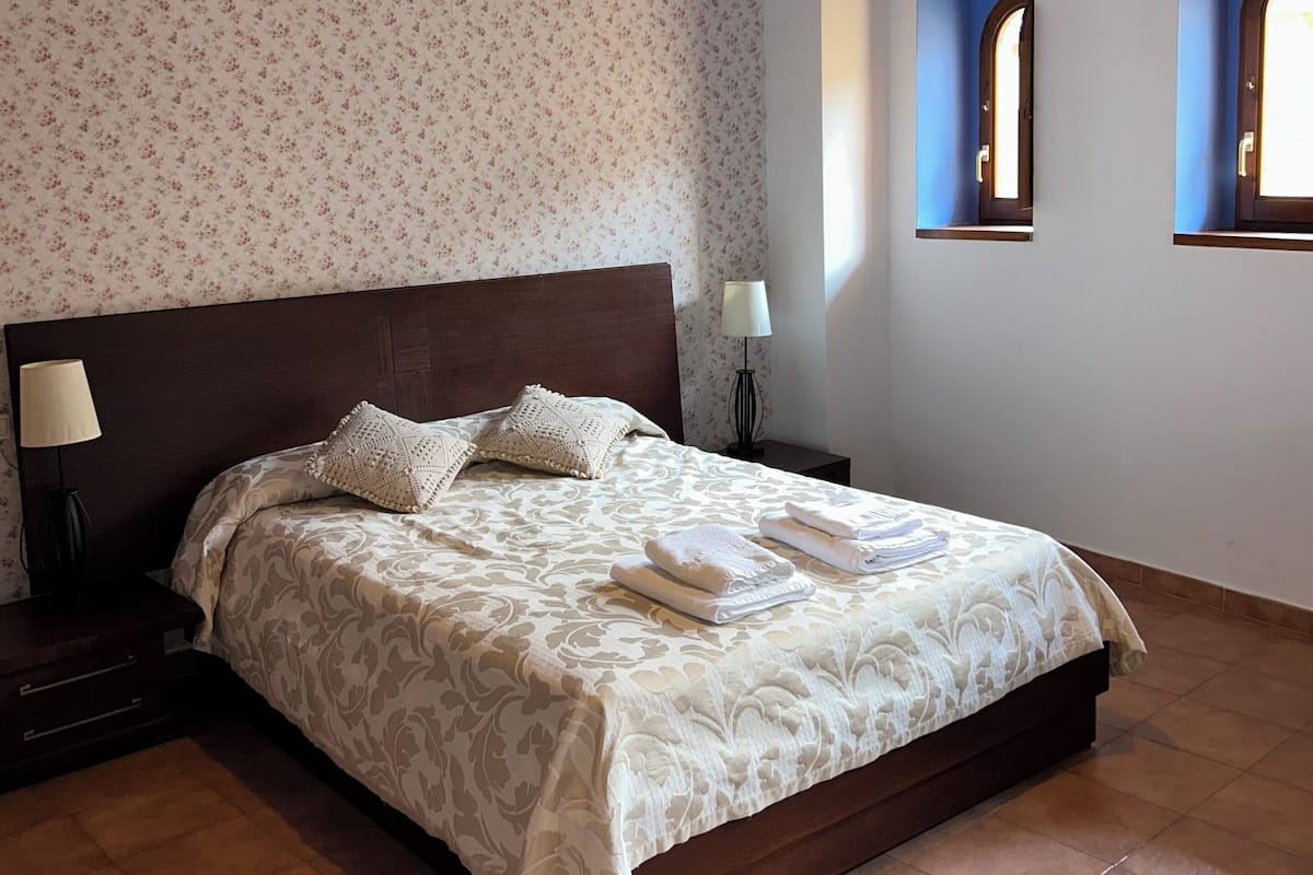 A bedroom with a large bed, wooden headboard, two bedside lamps, a hanging light fixture, and a window with a view.