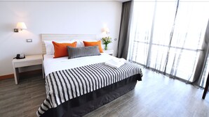 Ein modernes Hotelzimmer mit einem großen Bett, zwei Nachttischen, einem Fenster mit Vorhängen und Blick auf die Stadt.