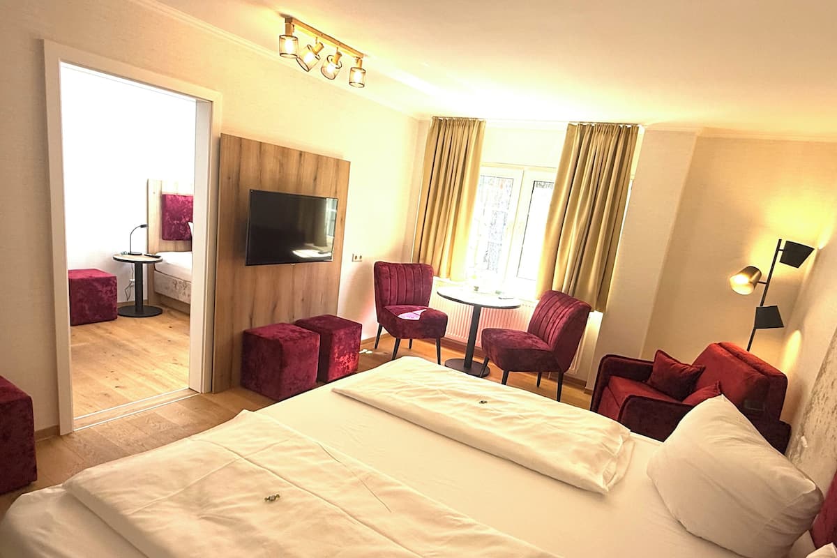 Une chambre d’hôtel avec un lit, deux fauteuils rouges, une petite table, un téléviseur et une fenêtre avec des rideaux.