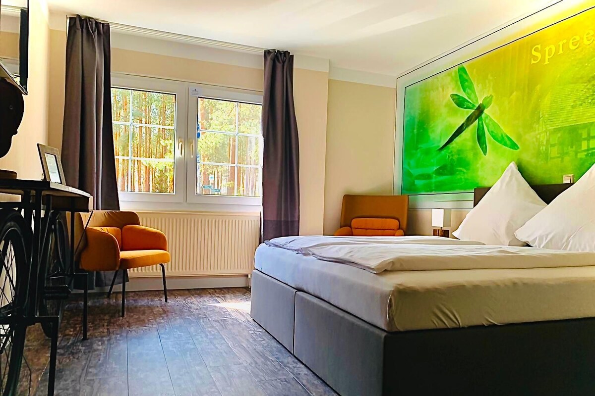 A hotel room with a bed, a desk with a chair, a large window with curtains, and a wall-mounted TV.