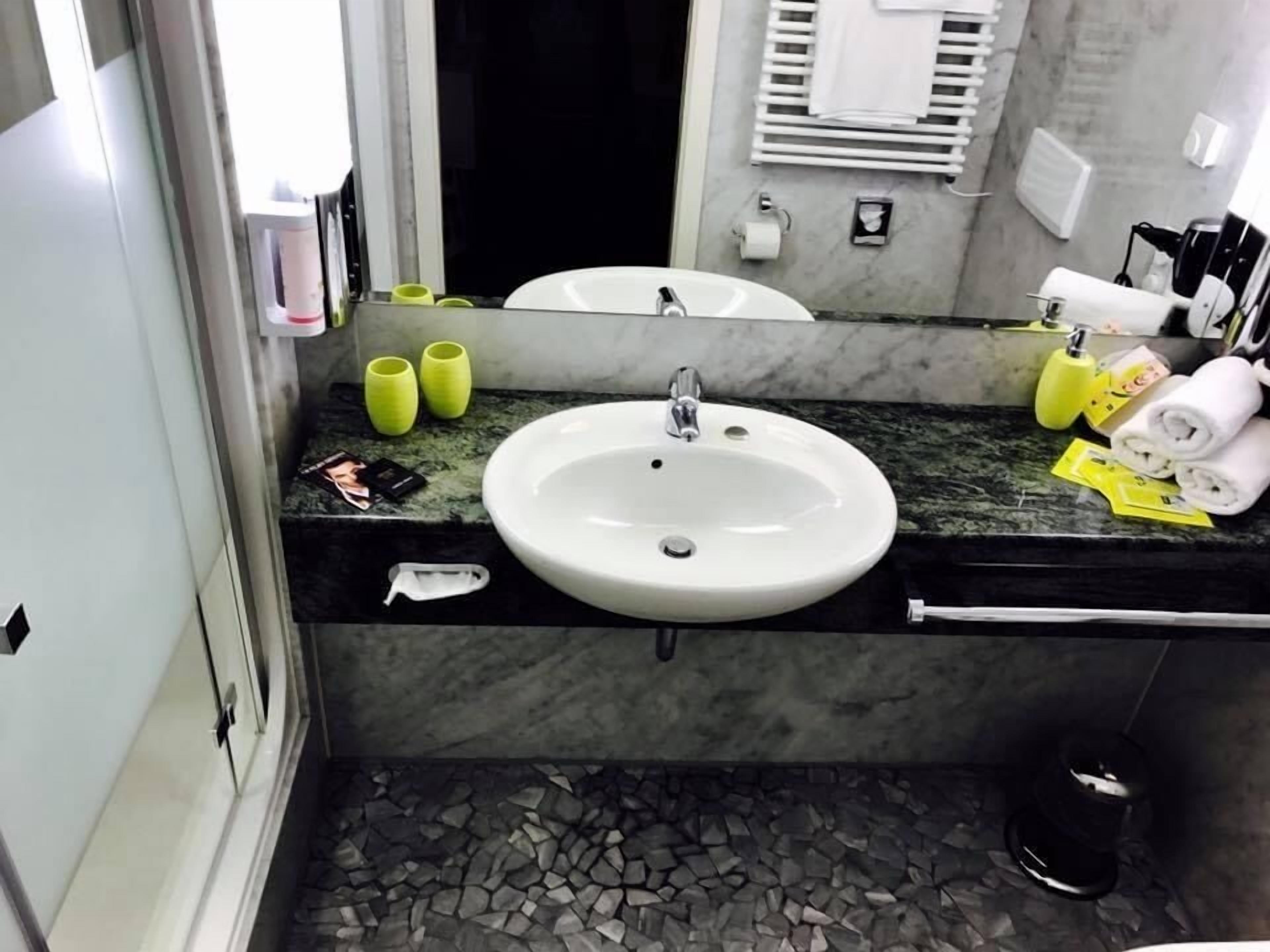 Een badkamer met een witte wastafel, een marmeren aanrechtblad en een spiegel. Handdoeken, toiletartikelen en een prullenbak zijn aanwezig.