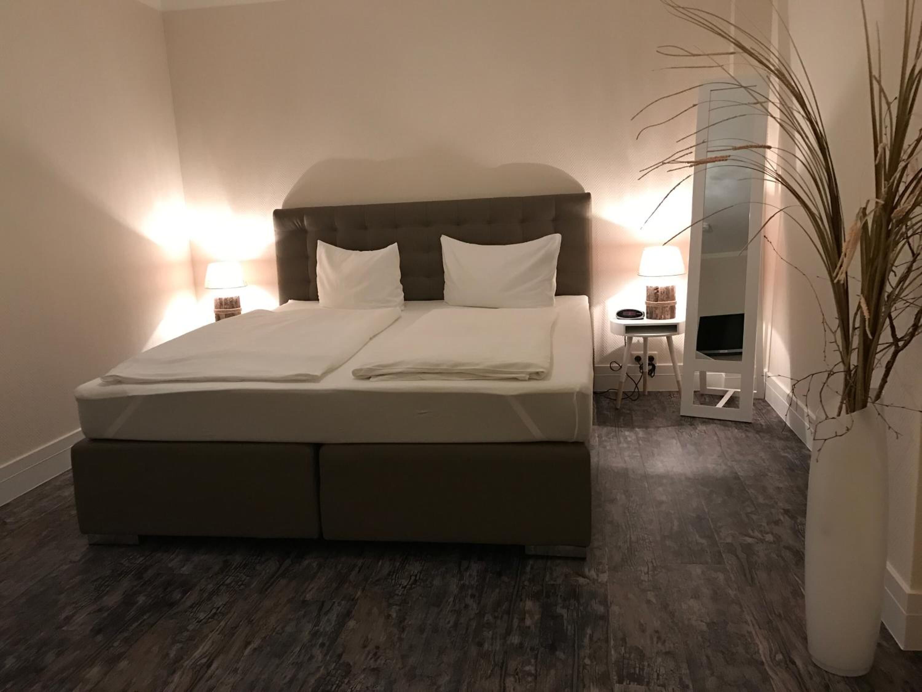 Una cama bien hecha con cabecera, dos lámparas de noche, una mesita con lámpara y un espejo grande en la pared.