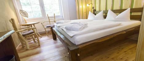 A wooden bed with pillows, a wooden chair, a small table, and a window with blinds.