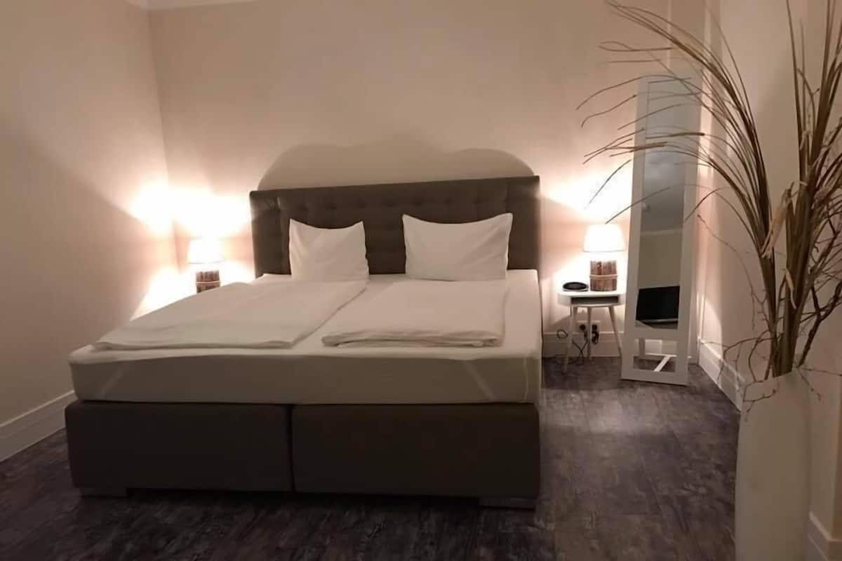 Un lit bien fait, avec une tête de lit, une table de chevet avec une lampe et un grand miroir au mur.