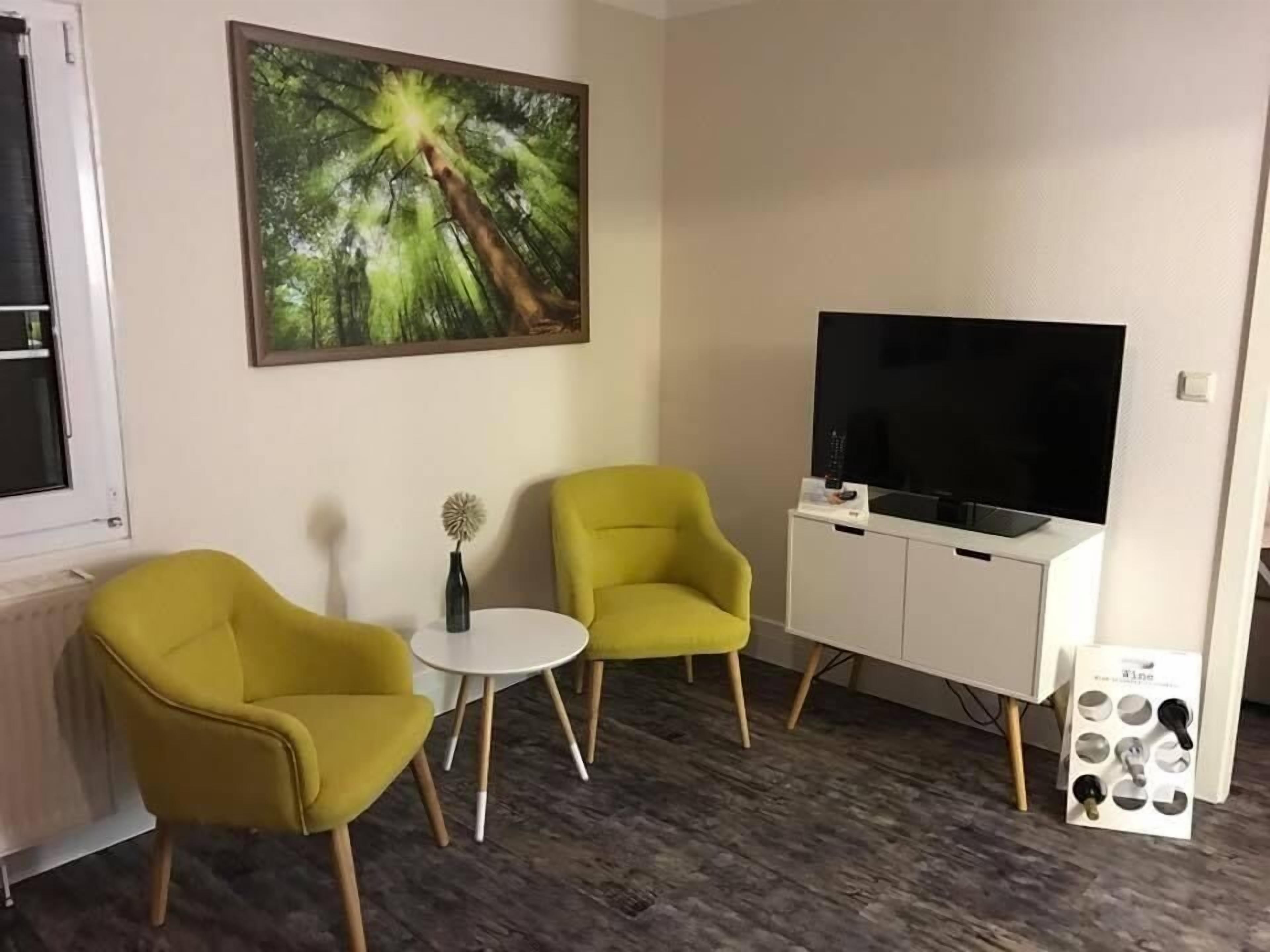 A room with two yellow armchairs, a small white table, a painting of a tree, a white cabinet with a TV, and a shoe rack.