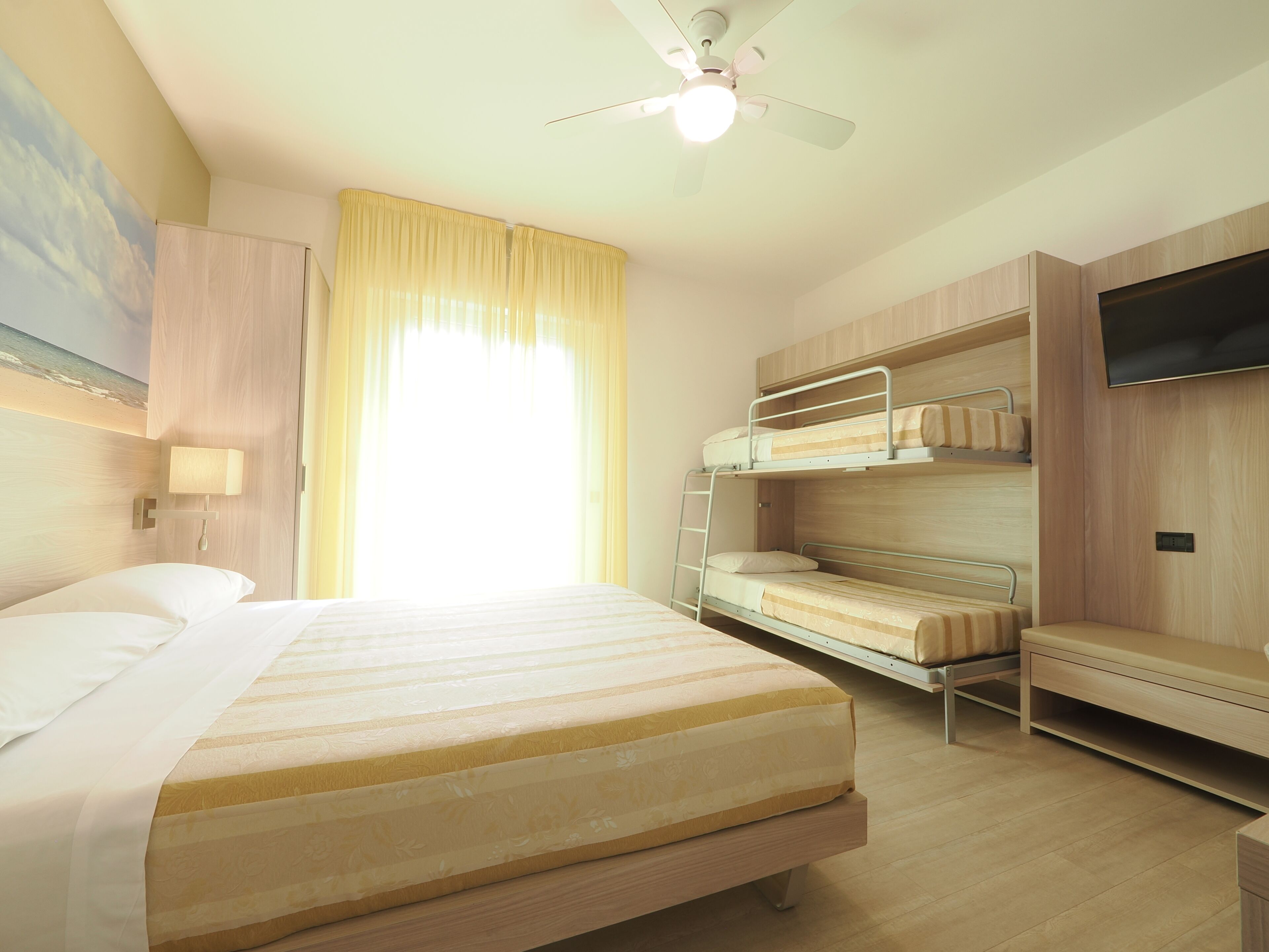 A hotel room with a bed, bunk beds, a ceiling fan, a TV, and a window with curtains.