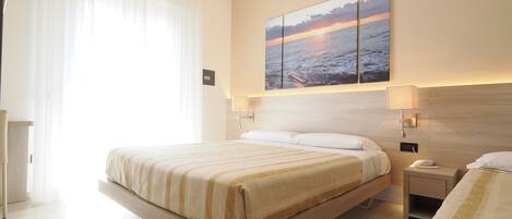 Une chambre d’hôtel moderne avec un grand lit, des tables de chevet, une lampe fixée au mur et un tableau représentant un coucher de soleil sur l’eau.