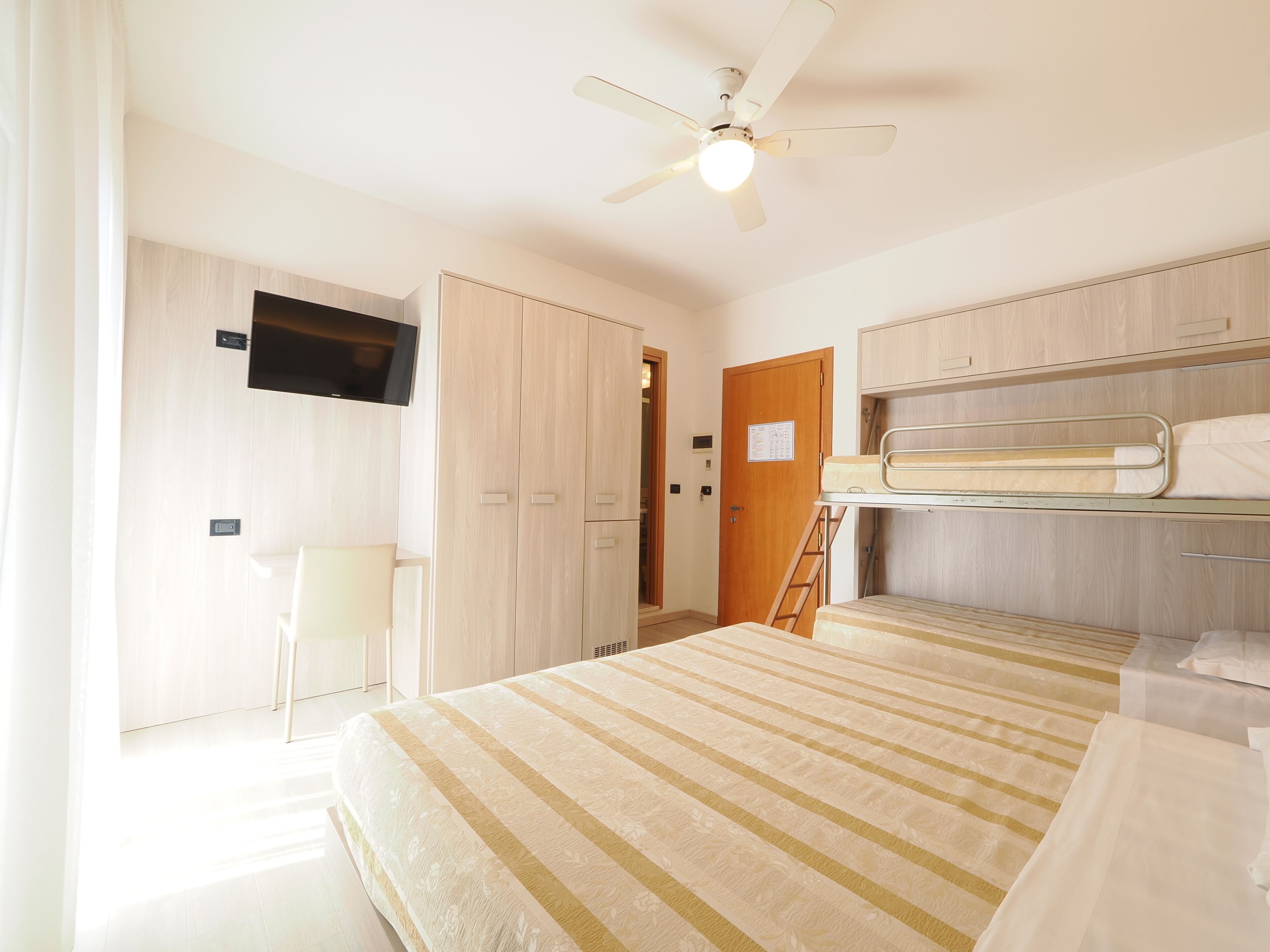 A bedroom with a bed, a desk, a chair, a wardrobe, a television, a ceiling fan, and bunk beds.
