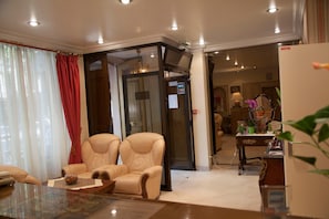 A hotel room with a seating area featuring two beige armchairs, a glass coffee table, and a red curtain. - Hôtel des Pyrénées - Nation (Paris)