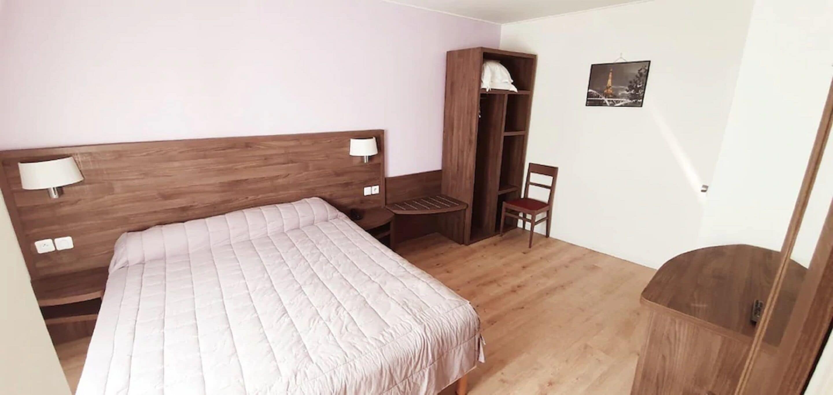 Un dormitorio con cabecera de madera, cama con colcha blanca, mesita de noche, armario de madera, una silla y estante fijado a la pared.