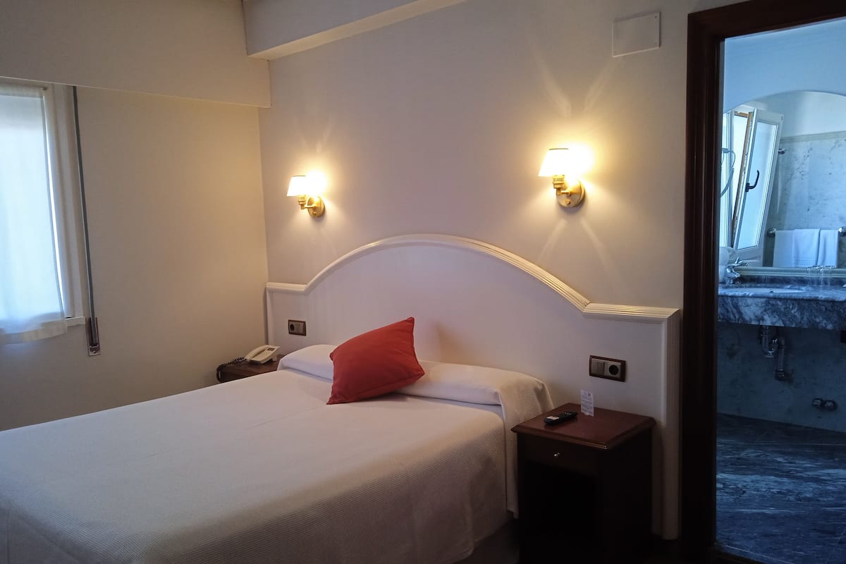 A hotel room with a bed, two bedside lamps, a nightstand, and a bathroom visible through an open door.