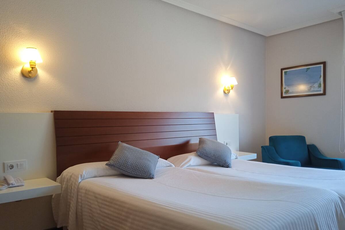 A hotel room with a large bed, two bedside lamps, a blue armchair, and a painting on the wall.