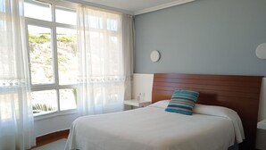 A bedroom with a bed, a nightstand, a wall clock, and a window with curtains. - Hotel Sablón (Llanes)