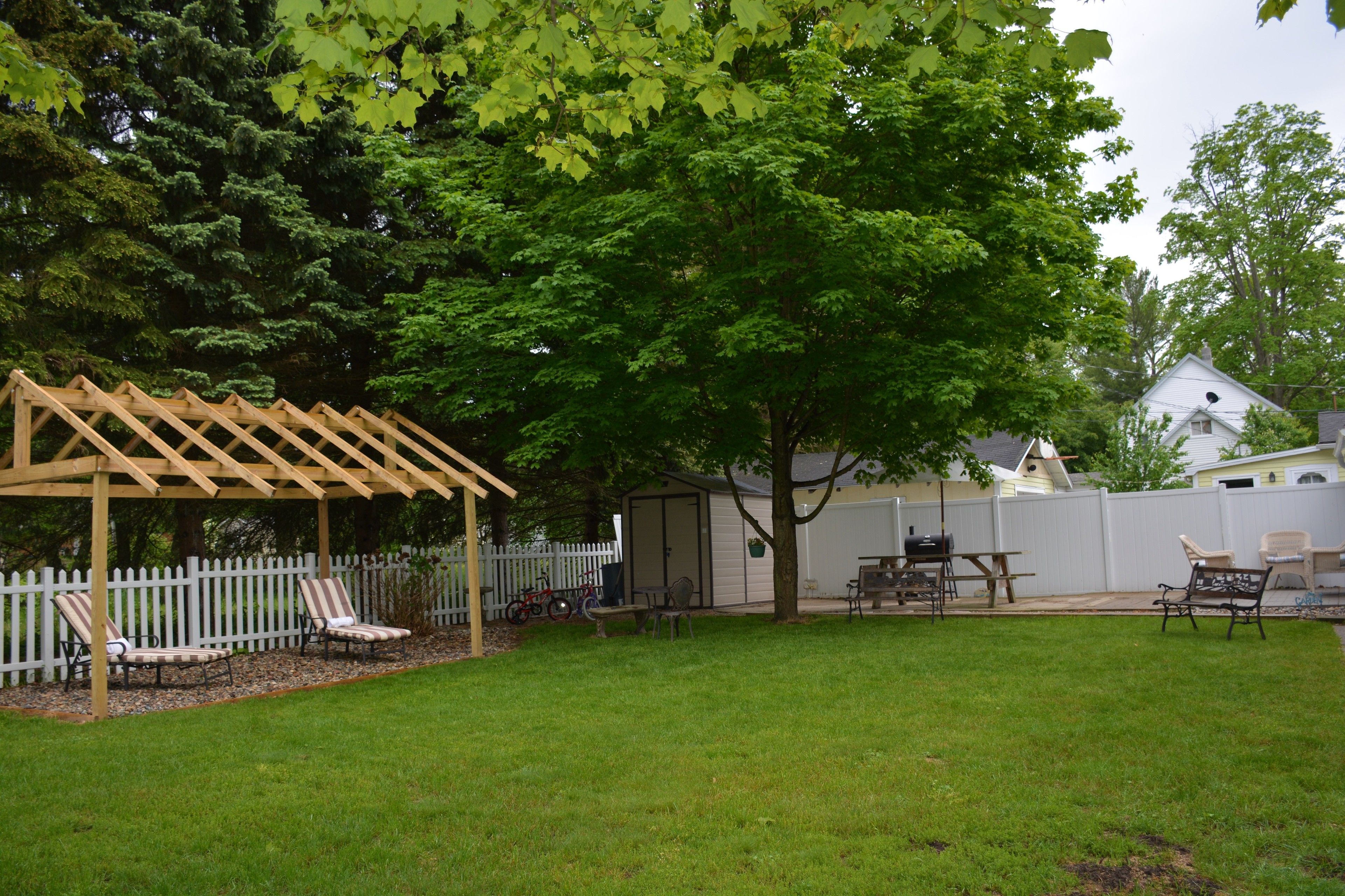 A backyard with a wooden pergola, two lawn chairs, a barbecue grill, and a shed.