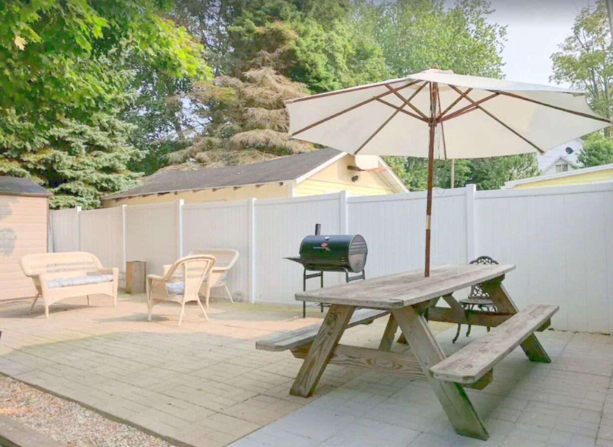 An outdoor patio with a wooden table and benches, a grill, and a white umbrella.