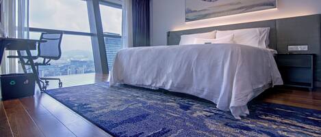 A modern hotel room with a large bed, a desk, and a view of the city through large windows.