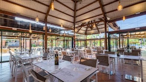 An outdoor dining area with white tables and chairs, hanging light fixtures, and large windows offering views of the surrounding greenery.