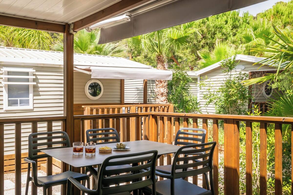 Une terrasse couverte, agrémentée d’une table et de chaises, entourée de rampes en bois et d’une végétation luxuriante.