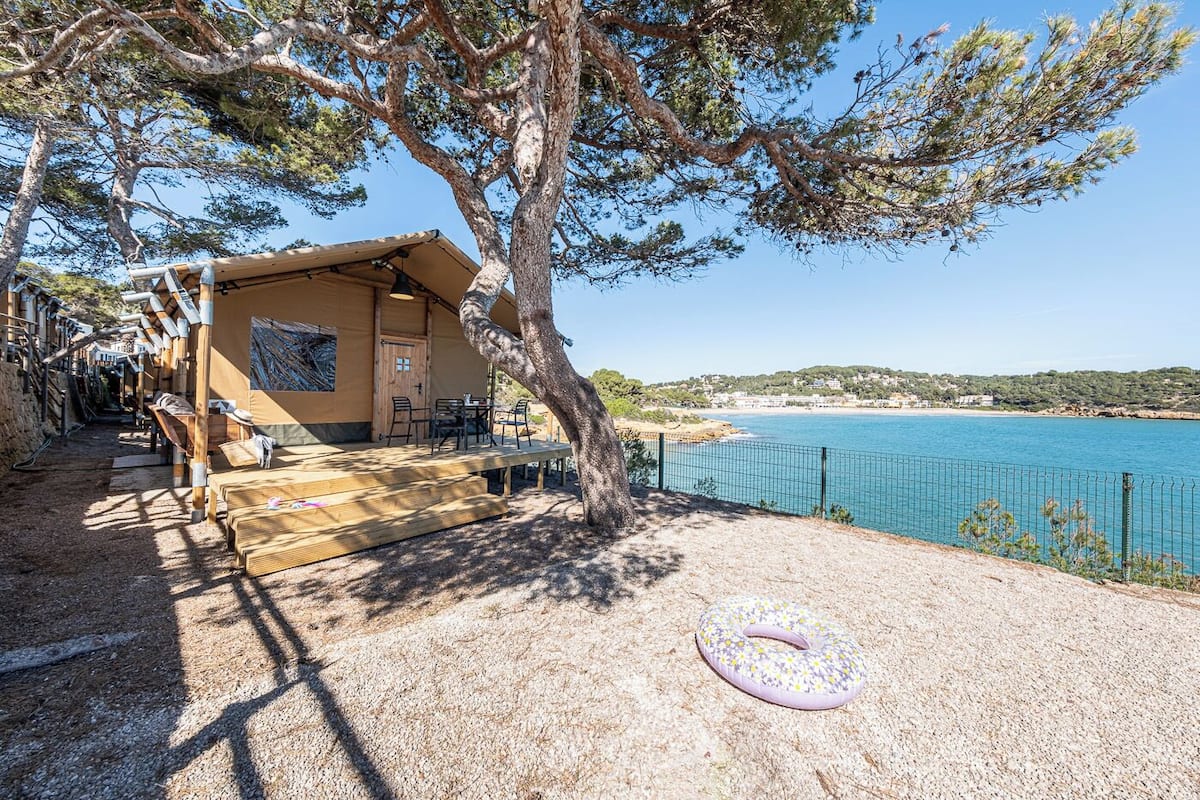 Une terrasse en bois avec des sièges extérieurs, un petit bâtiment doté d’une porte et de fenêtres, et une vue sur une étendue d’eau bleue.