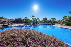 A clear blue pool surrounded by palm trees and purple flowers, with a wooden deck and a building in the background. - Estival Torre de la Mora (Tarragona)