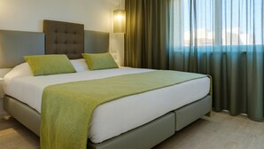 Ein modernes Hotelzimmer mit einem großen Bett, grünen Kissen, einem grauen Kopfteil und einem Fenster mit Vorhängen.