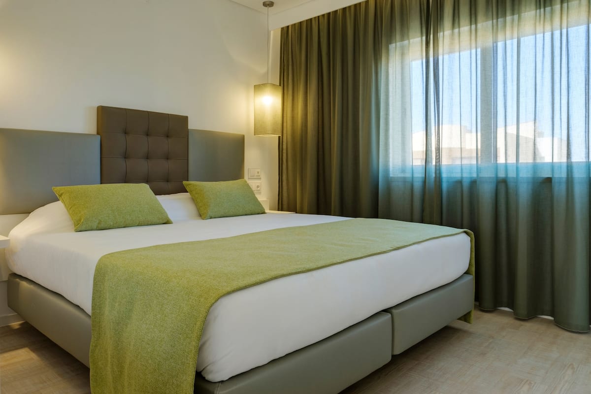 Una moderna camera d'albergo con un letto grande, cuscini verdi, una testiera grigia e una finestra con tende.