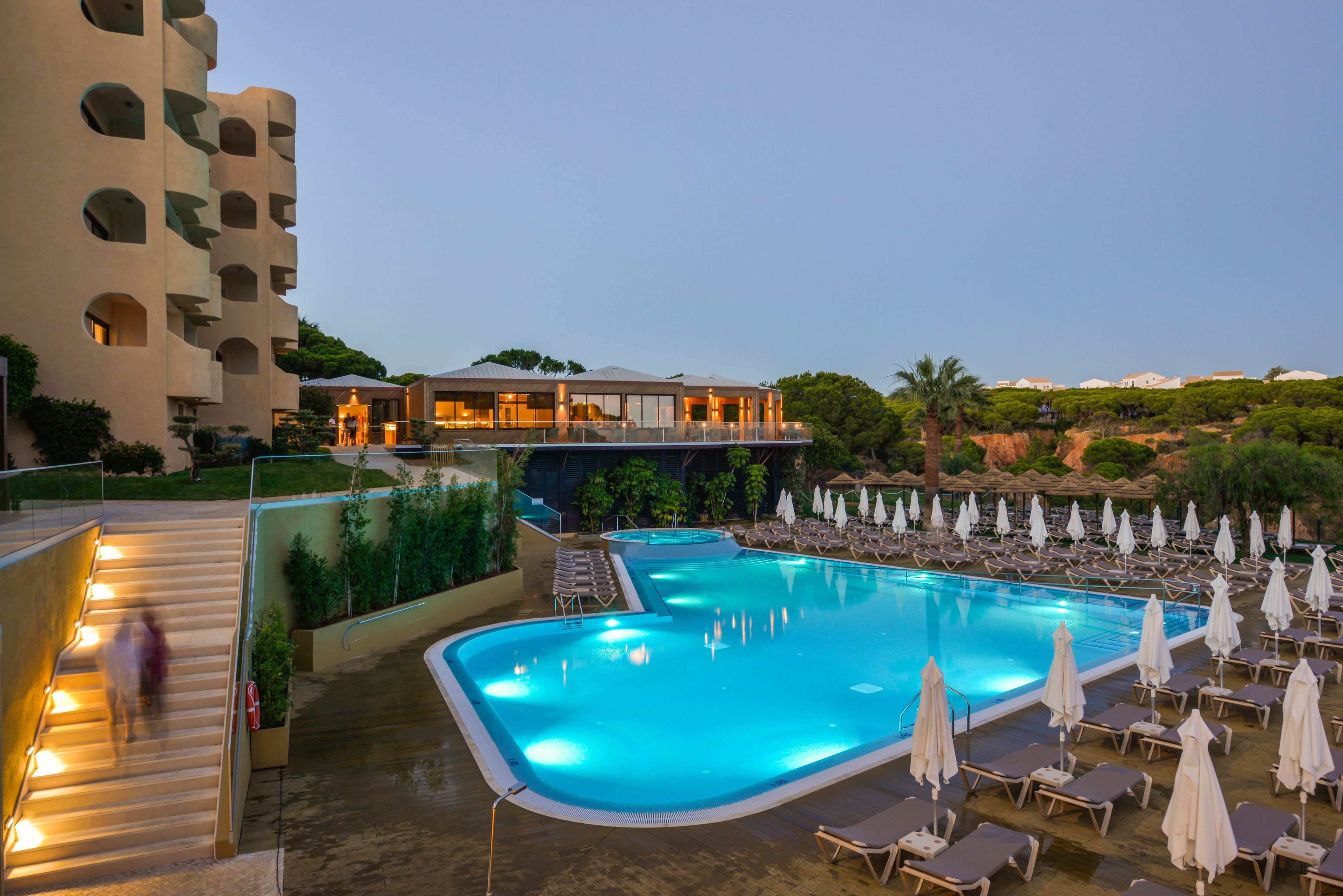 Una piscina con sillones de descanso y sombrillas, una escalera y un edificio con varios balcones.