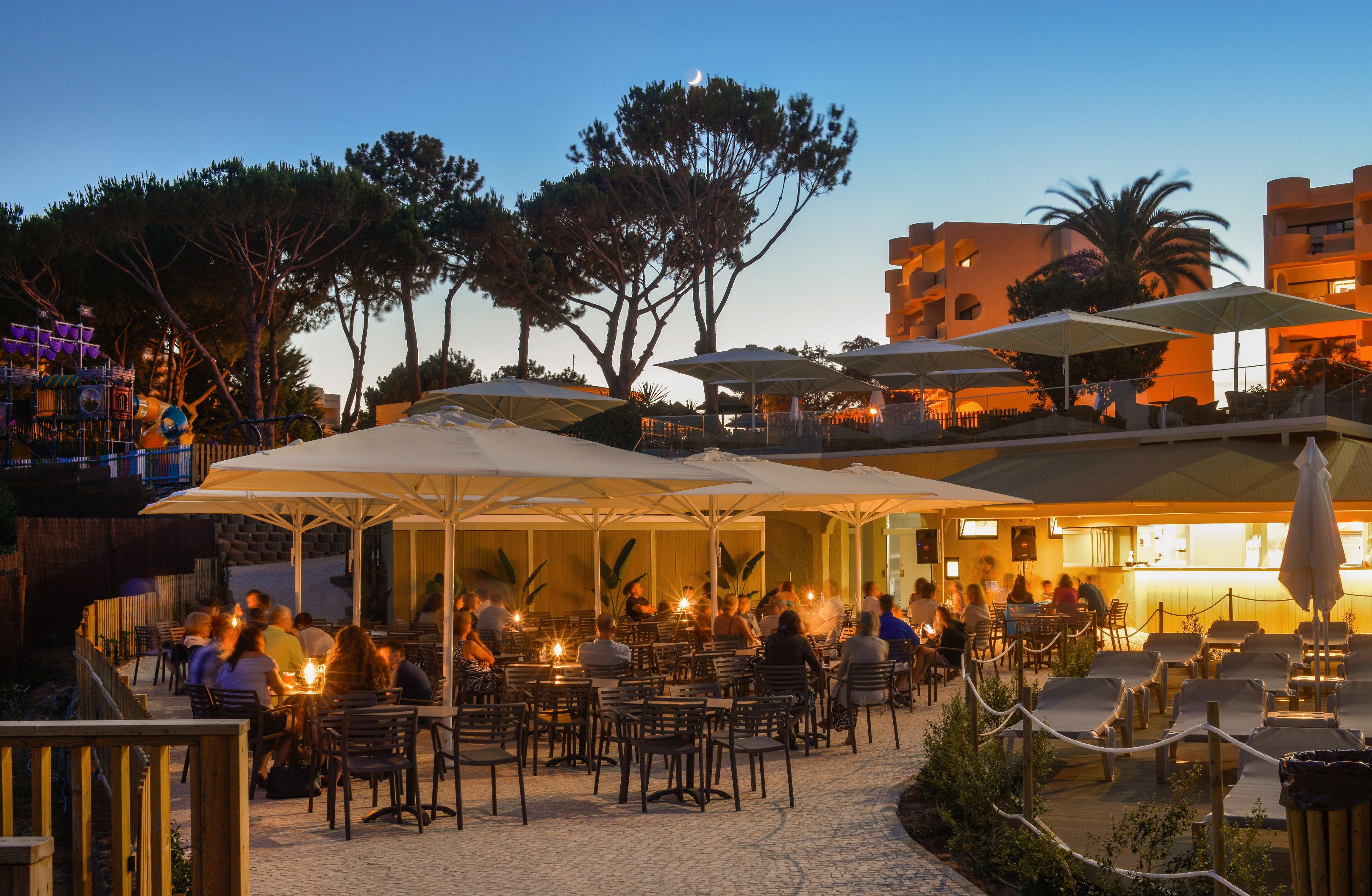 Un espace extérieur aménagé pour les repas, avec des parasols, des tables et des chaises, entouré d’arbres et de bâtiments.
