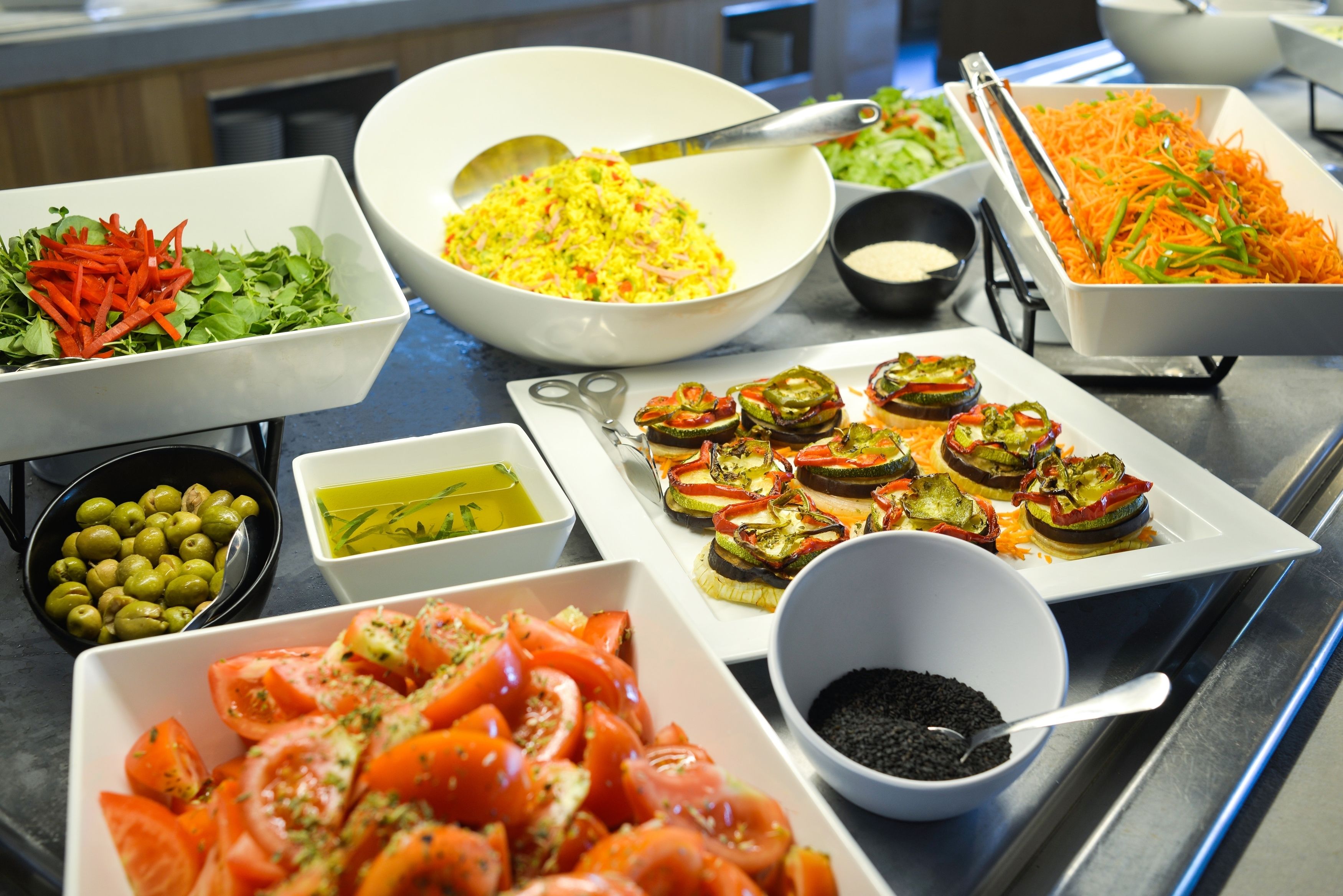 Une table de buffet proposant divers plats, notamment un bol de riz, une assiette de tomates en tranches, un bol d’olives et un plateau de légumes coupés en tranches.