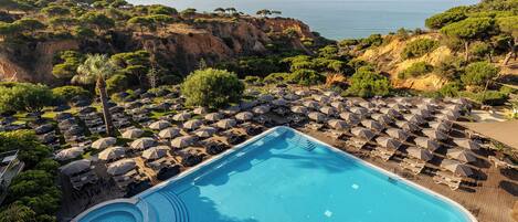 Un espace piscine doté de chaises longues et de parasols, entouré d’arbres et offrant une vue sur une falaise.