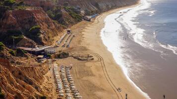 A coastal beach with rows of sunbeds and umbrellas, a sandy beach, and a cliffside.
