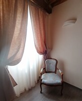 Una stanza con una sedia vicino a una finestra con tende leggere, una parete semplice e un pavimento in legno.
