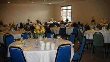 Uma sala de banquetes com mesas redondas preparadas para refeições, cada uma com toalhas brancas, cadeiras azuis e guardanapos amarelos.